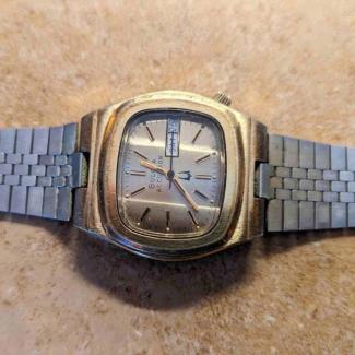Gold, square-faced watch with Bulova Accutron text, tuning fork symbol, and date/day window