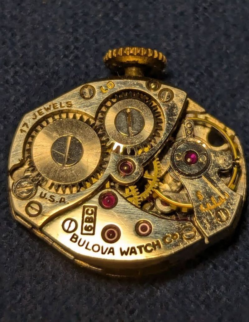 A close picture of the Bulova movement. The crown is at the top of the picture.  Directly below the crown is the engraving L0. To the left of that is engraved "17 JEWELS." Opposite of the crown, along the edge is engraved "BULOVA WATCH Co" and there is a small box perpendicular to that with the engraving 6BC. There is also a U.S.A engraving to the left, and to the right there is the regulator pin and the engraving 5 